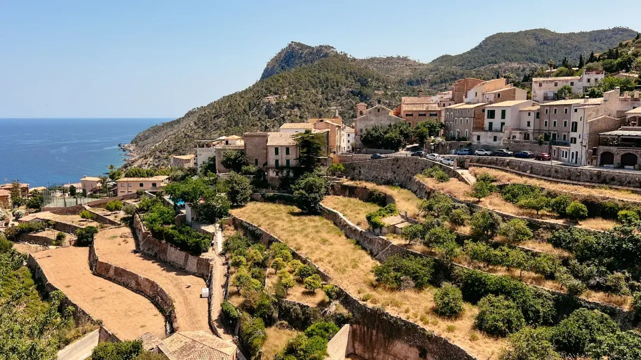 Terrassierte Hänge mit Bäumen und trockener Vegetation, die sich zum Meer hin erstrecken. Im Hintergrund liegt ein Dorf am Fuße des Tramuntana Gebirges.