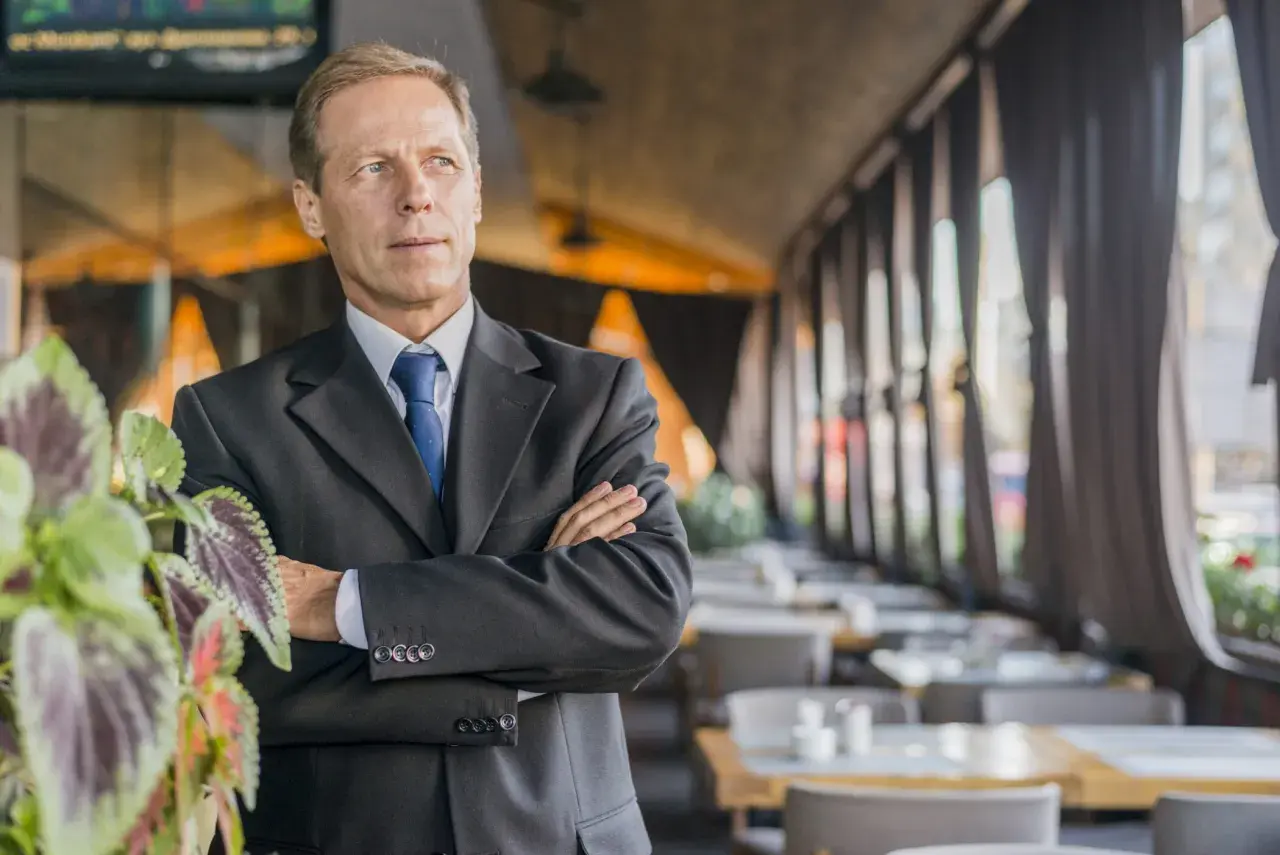 Uomo d'affari con braccia conserte, un chief people officer che guarda lontano, con piante verdi in primo piano e un ristorante sullo sfondo.
