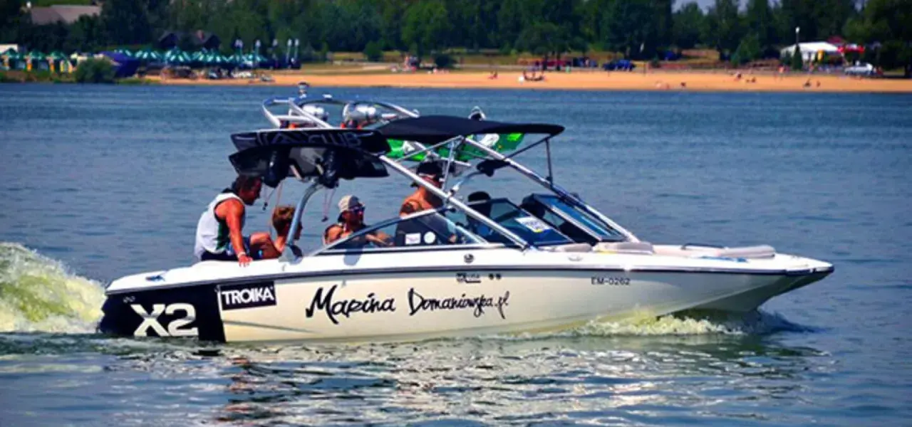 Łódź motorowa z pasażerami płynie po wodzie, tworząc pianę. Na burcie widnieje napis "Marina Domaniewska".