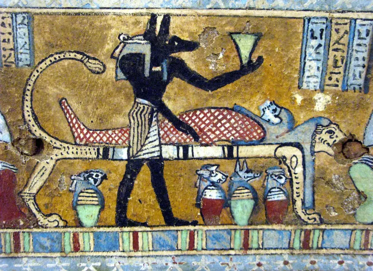 Anubis, bóg z głową szakala, stoi przy katafalku z mumią. W ręku trzyma naczynie, a obok stoją urny z głowami zwierząt.