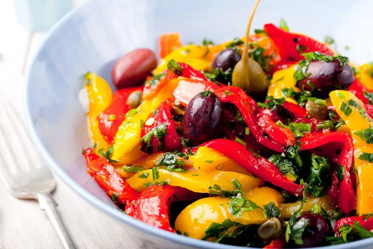 Insalata colorata di peperoni in agrodolce, con olive nere, capperi e prezzemolo fresco, servita in una ciotola blu.