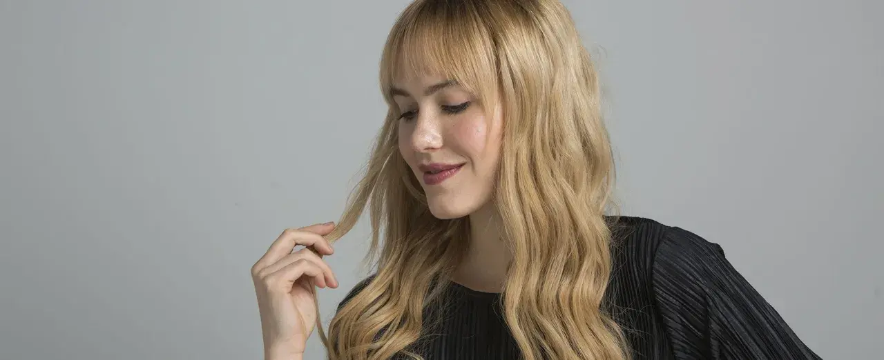 Mujer con cabello rubio ondulado y flequillo, tocando un mechón. El matizar el pelo realza su brillo y tono natural.