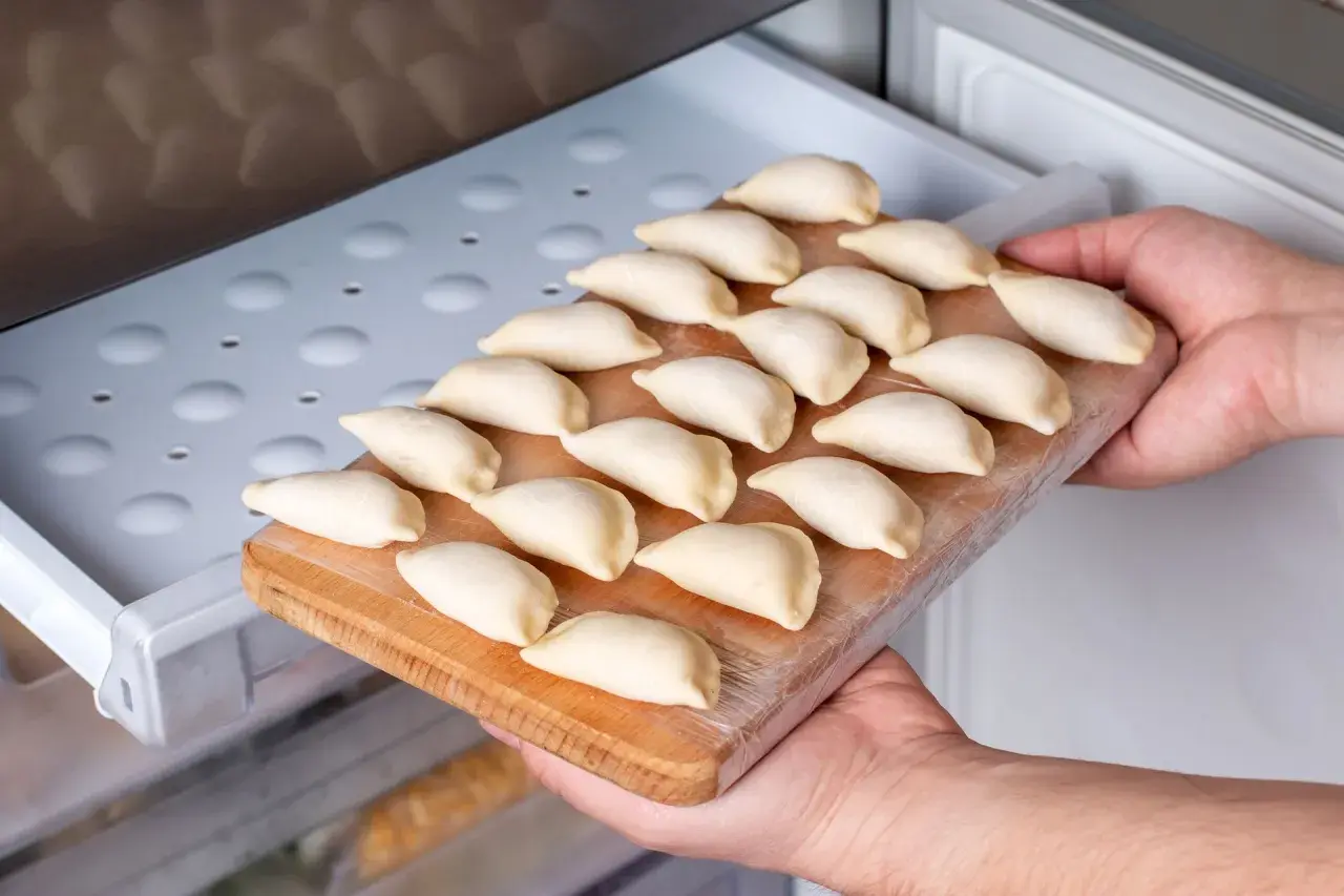 Ręce wkładają deskę z surowymi pierogami do zamrażarki. To prosty sposób, jak przechować ciasto na pierogi na później.