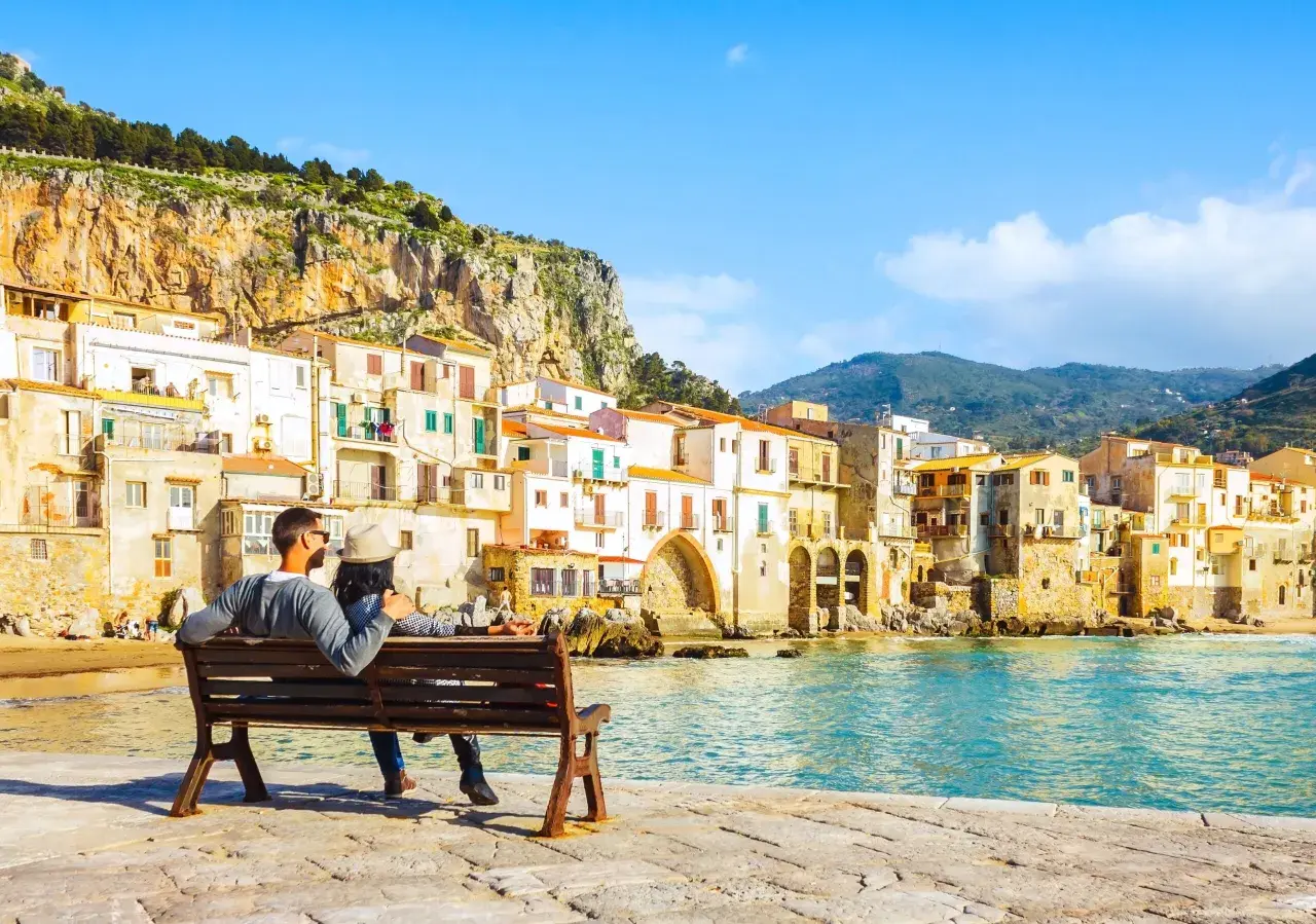 Coppia seduta su una panchina, ammira il mare e le case colorate di Cefalù, un luogo perfetto per vacanze in Sicilia sul mare.