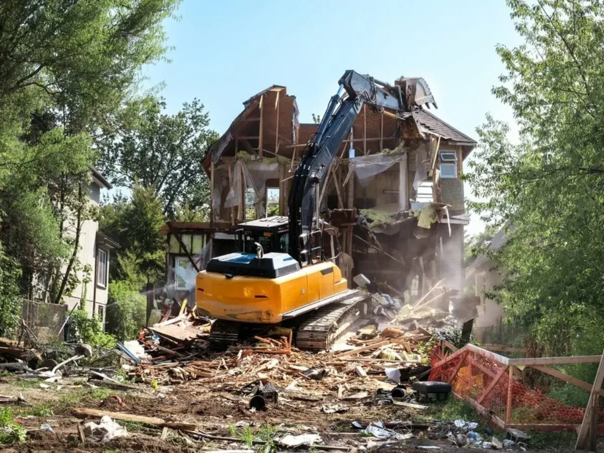 Żółty koparka prowadzi rozbiórkę starego domu, zasypując teren gruzem.