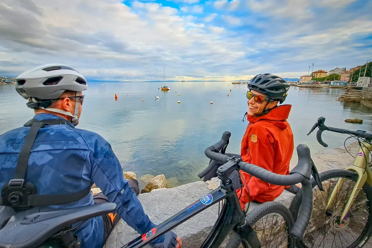 Two cyclists enjoy a break during bike guided tours in the Mediterranean, with boats on the calm sea and a coastal town in the background.
