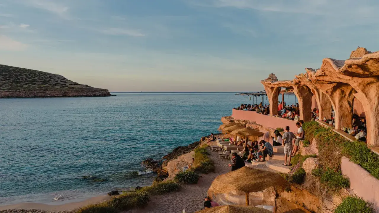 Menschen genießen den Sonnenuntergang am Strand von Ibiza, in der Nähe eines einzigartigen ashram-ähnlichen Gebäudes am Meer.