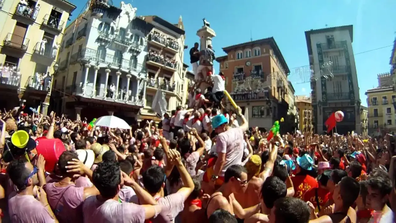 Multitud celebra cuando son vaquillas en Teruel. Gente en una plaza con edificios históricos, algunos subidos a un monumento.