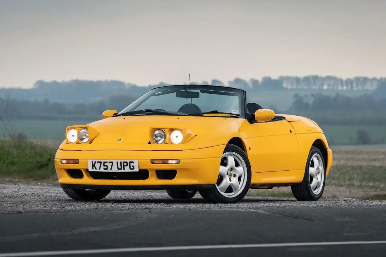 Gul Lotus Elan M100 cabriolet parkerad på grusväg med dimmig bakgrund.