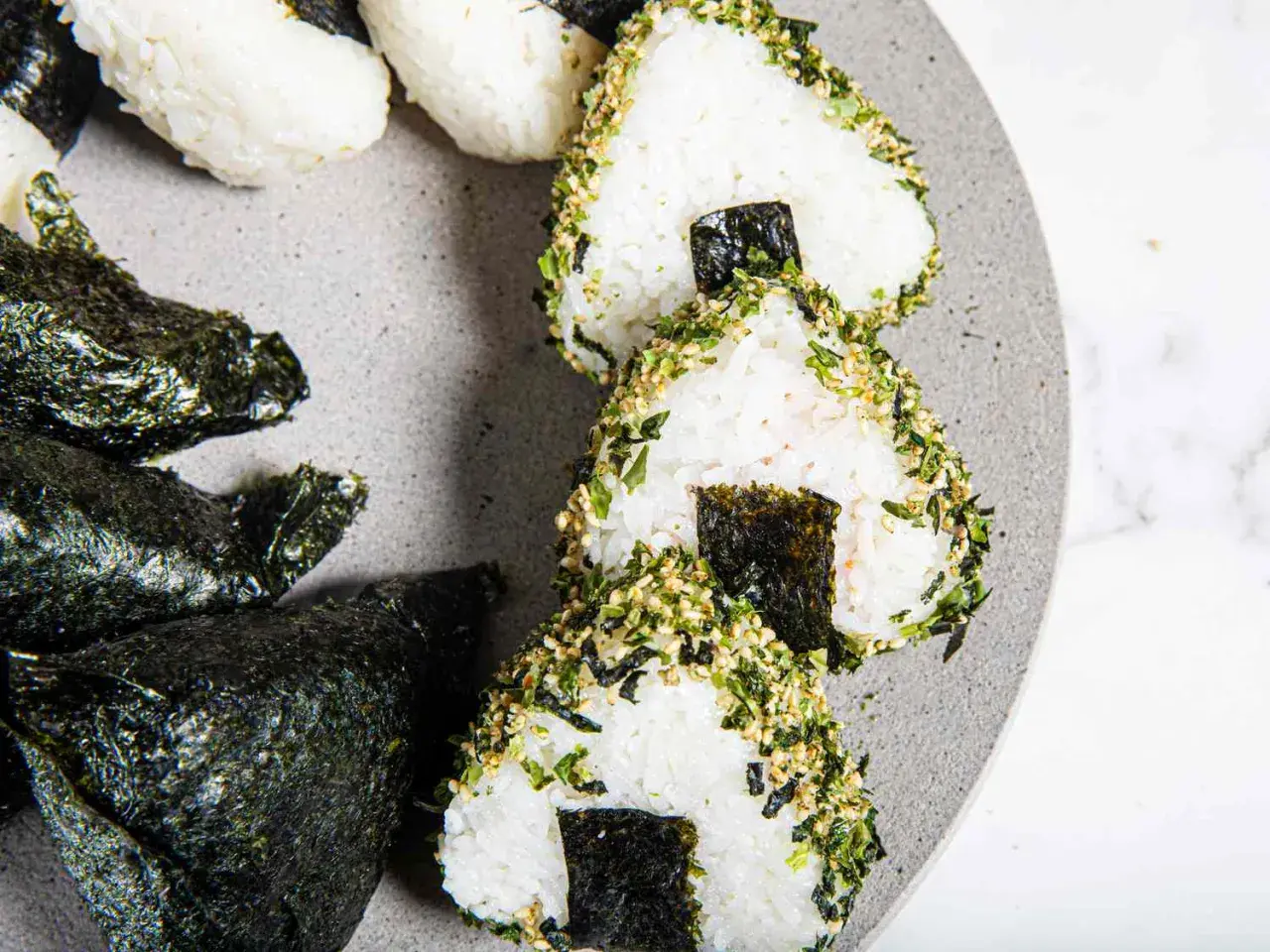 Triangular sushi rice balls, some wrapped in nori, others coated in sesame seeds and herbs, are artfully arranged on a grey plate.