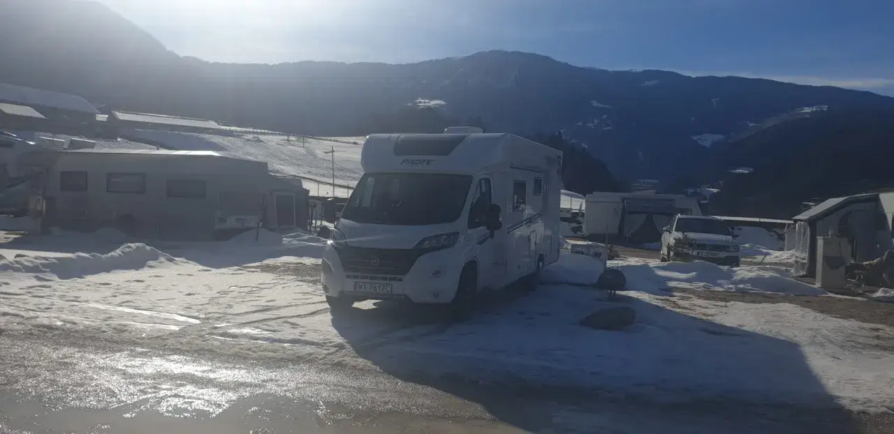 Kampery i przyczepy kempingowe na śniegu, gotowe na całoroczny camping w górach. Słońce przebija się przez chmury, oświetlając zimowy krajobraz.