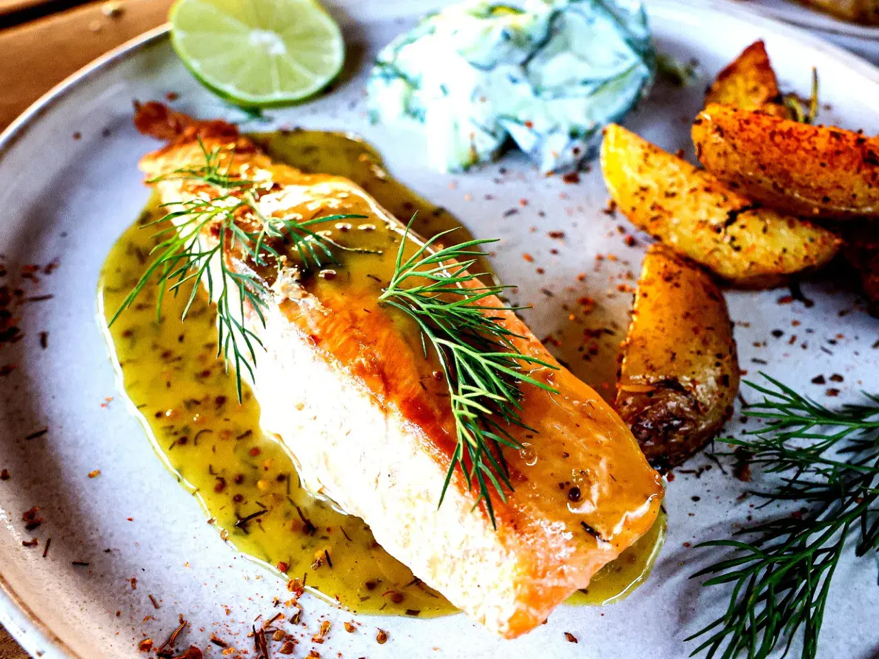 Gebratener Lachs mit Kräuterdressing, Kartoffelspalten und einem Klecks Gurkensalat. Ein warmes Gericht, das an Gurken Rezepte warm erinnert.