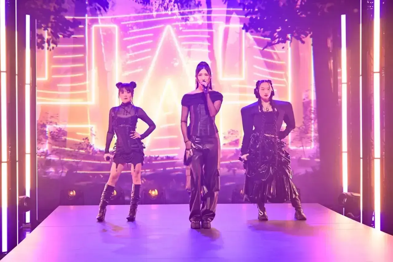 Trzy artystki k-pop w czarnych, stylizowanych strojach na scenie z neonowym tłem.