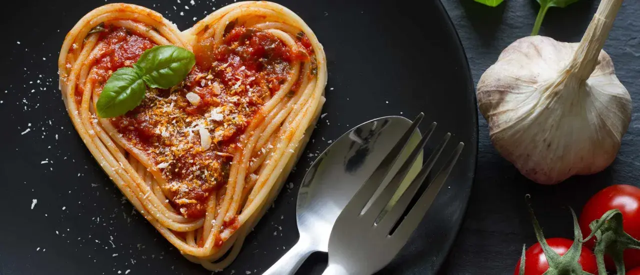 Herzförmige Pasta-Gerichte mit Tomatensoße, Basilikum und Parmesan. Daneben liegen ein Löffel, eine Gabel, Knoblauch und Tomaten.