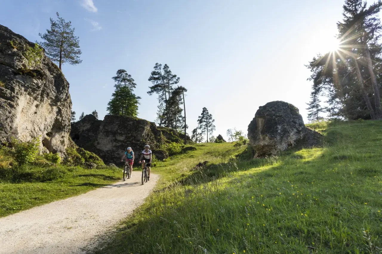 Zwei Radfahrer genießen eine Tour durch eine felsige Landschaft. Sie fahren auf einem Schotterweg, flankiert von grünen Wiesen und imposanten Felsen. Ein perfekter Tag, um in Heubach zu wandern.