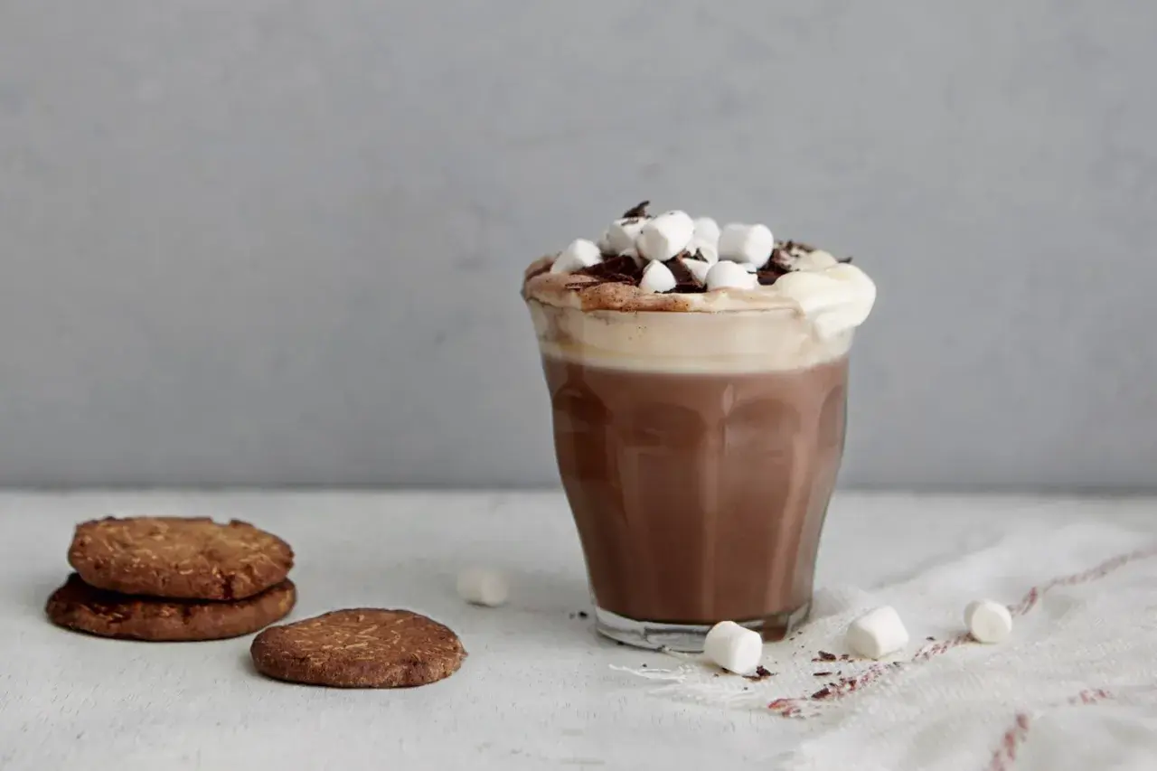 En härlig varm choklad med fluffigt skum, marshmallows och chokladströssel. Bredvid ligger några krispiga kakor.