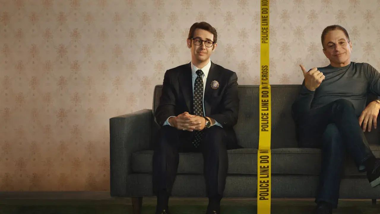 Dwóch mężczyzn siedzi na kanapie. Jeden, ubrany w garnitur, to **the good cop**. Obok niego, za taśmą policyjną, drugi mężczyzna uśmiecha się.