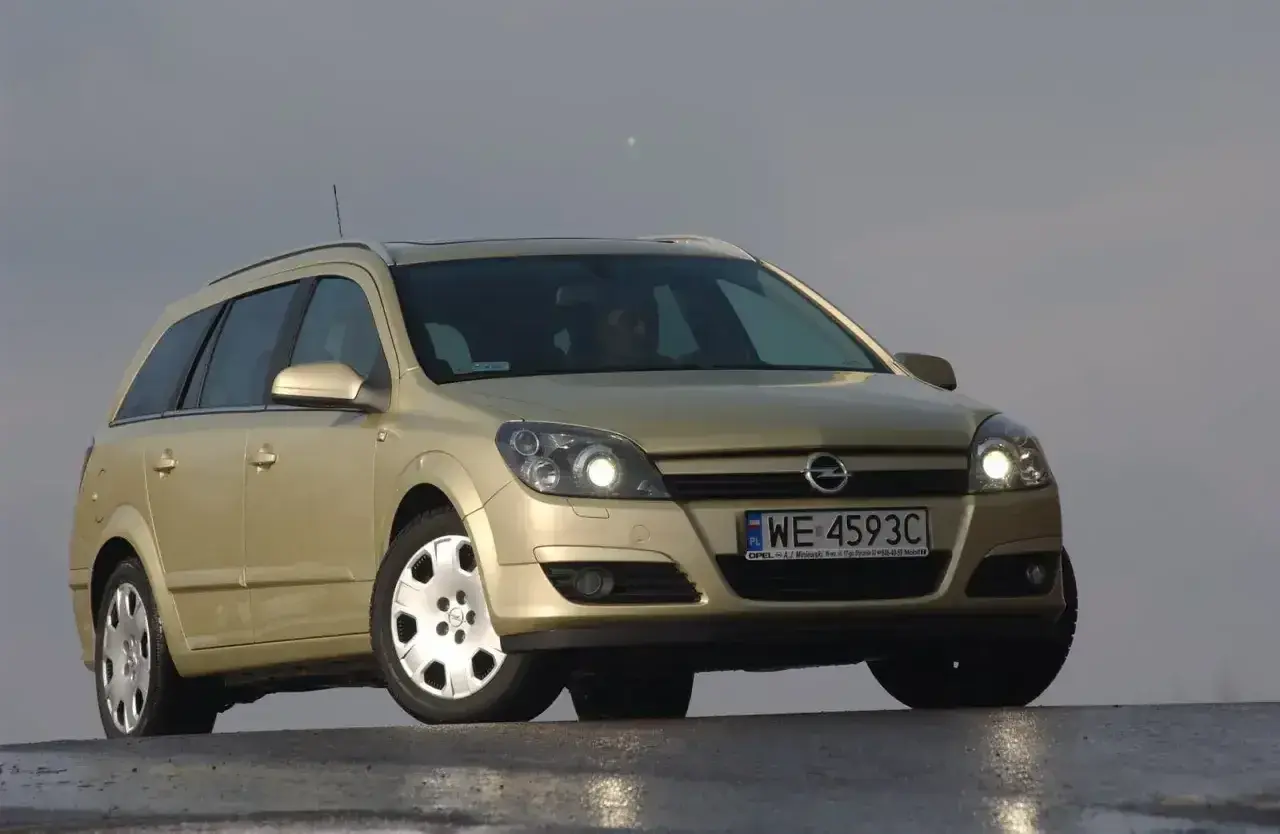 Złoty Opel Astra H kombi z włączonymi światłami. Przód samochodu z wyraźnie widocznymi żarówkami.