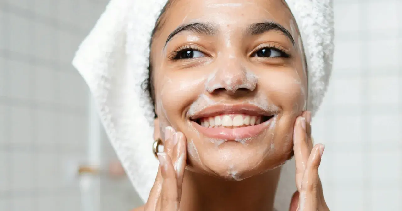 Una mujer sonríe mientras se aplica espuma en el rostro, demostrando en qué consiste la doble limpieza facial para una piel radiante.