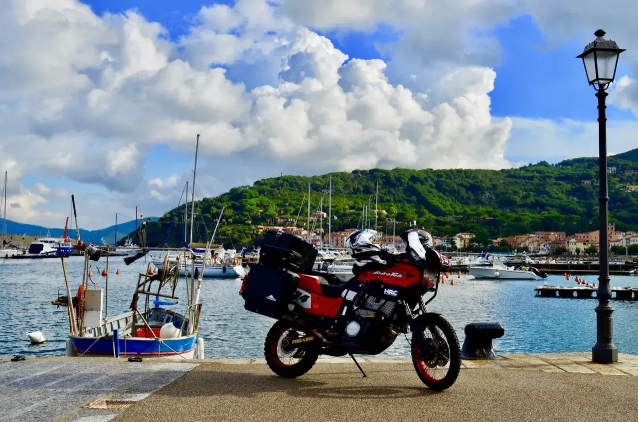 Moto rossa pronta a partire dal porto azzurro dell'Elba, con barche a vela e colline verdi sullo sfondo.