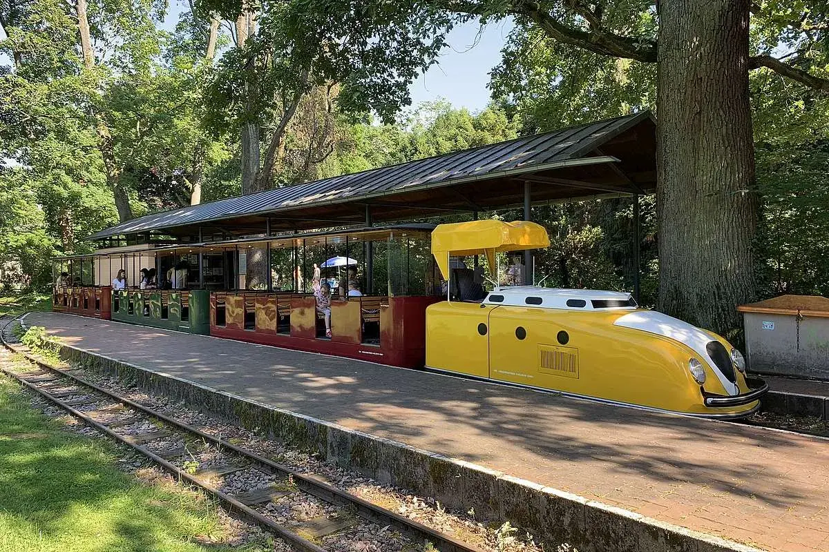Die gelbe Lokomotive der Schlossbahn Karlsruhe steht bereit. Fahrgäste sitzen in den roten und grünen Waggons unter einem schattigen Dach.