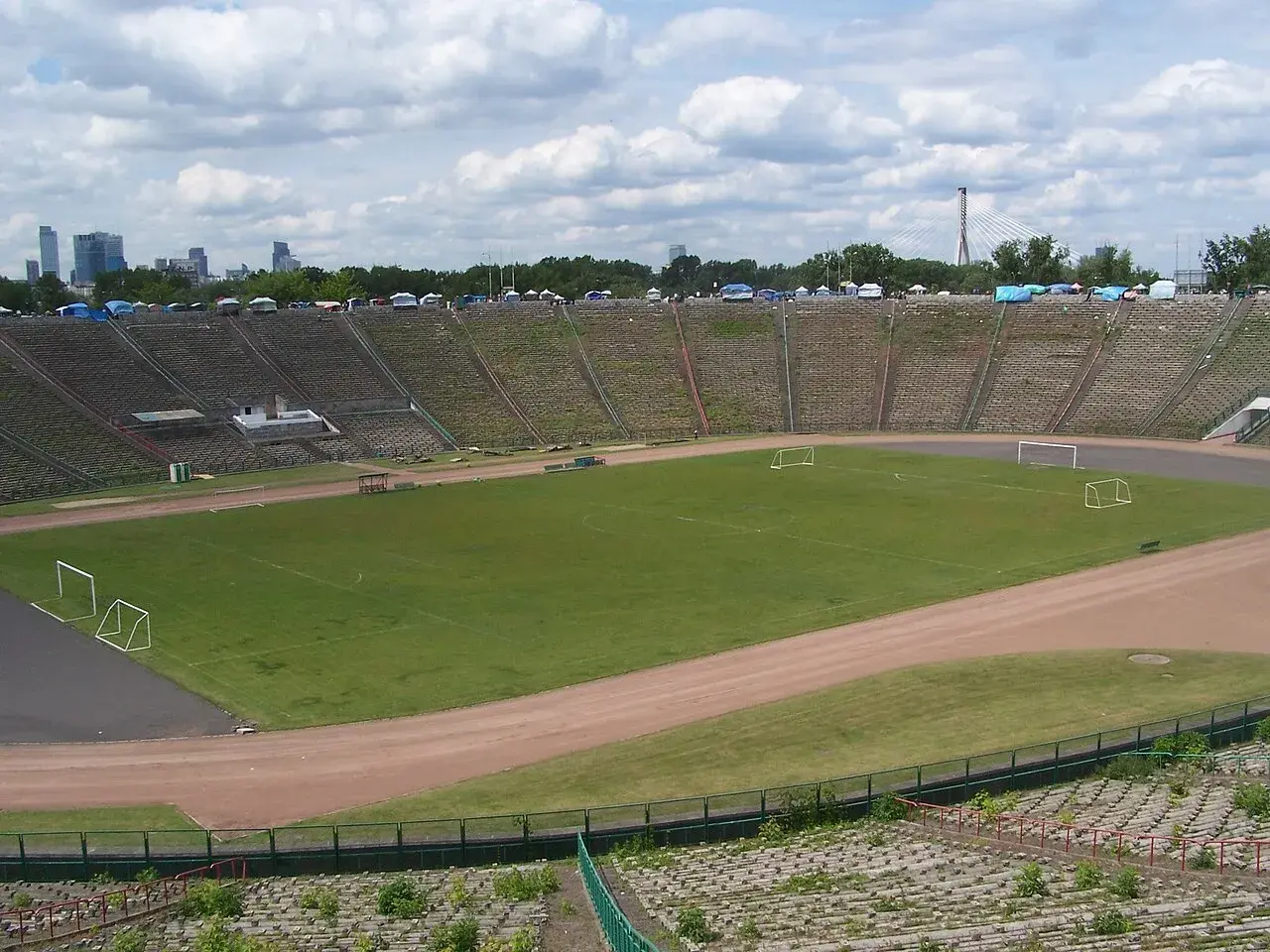 Stadion z PRL z murawą, bramkami i trybunami, w tle panorama Warszawy.