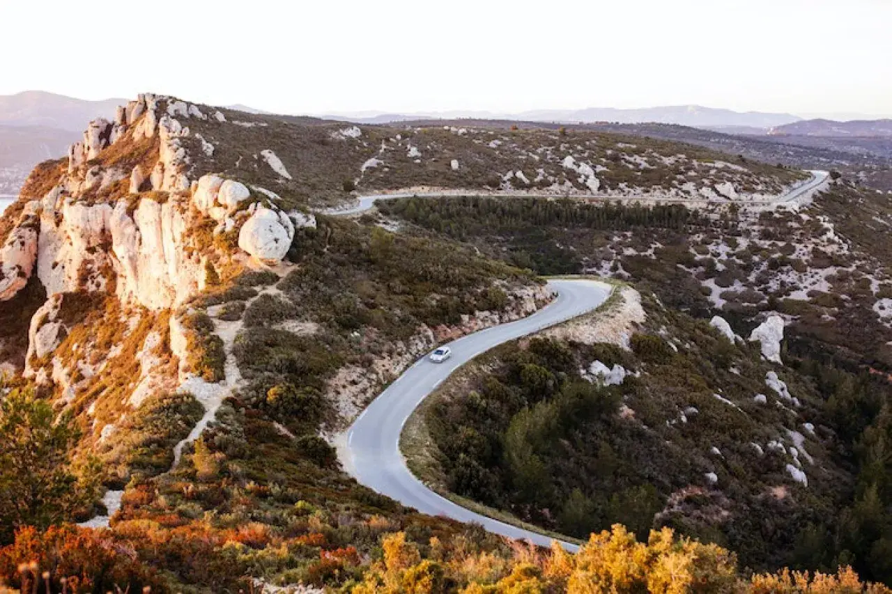 Route sinueuse à travers le massif du Cap Canaille, baignée par la lumière dorée du soleil couchant. Une voiture blanche serpente sur l'asphalte.