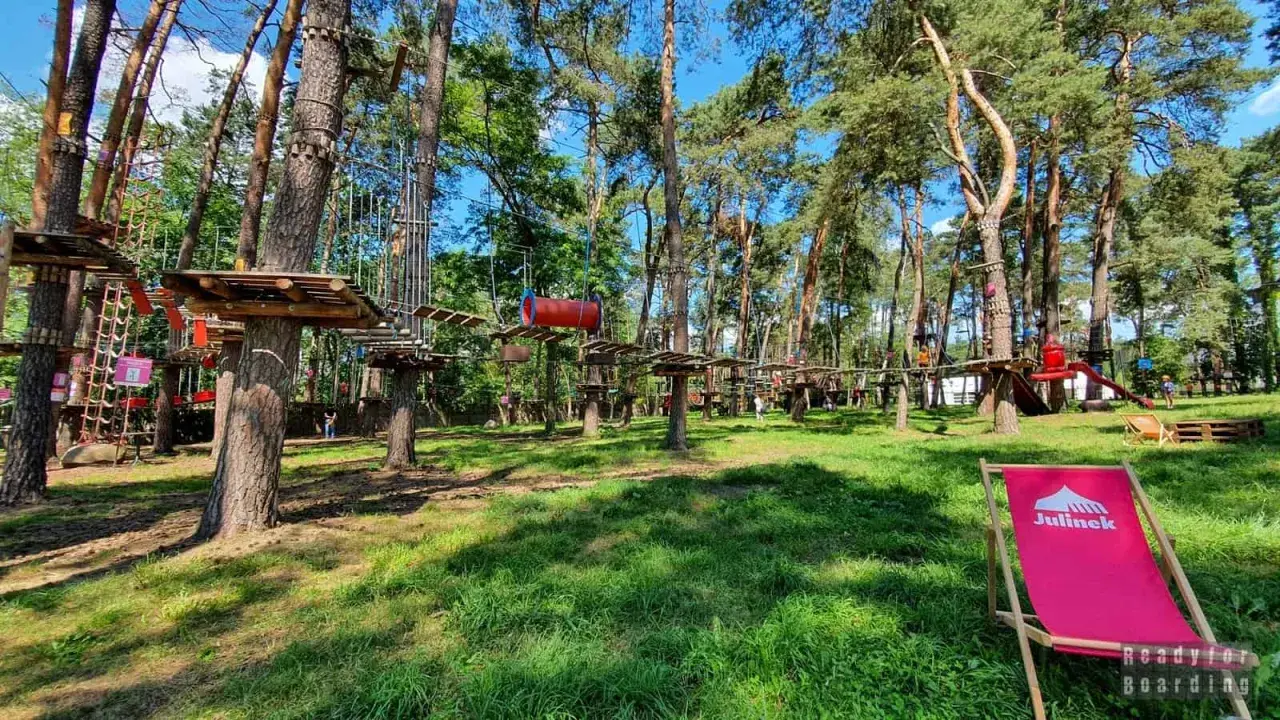 Park linowy w lesie z drewnianymi platformami i przeszkodami na drzewach. Na pierwszym planie różowy leżak.