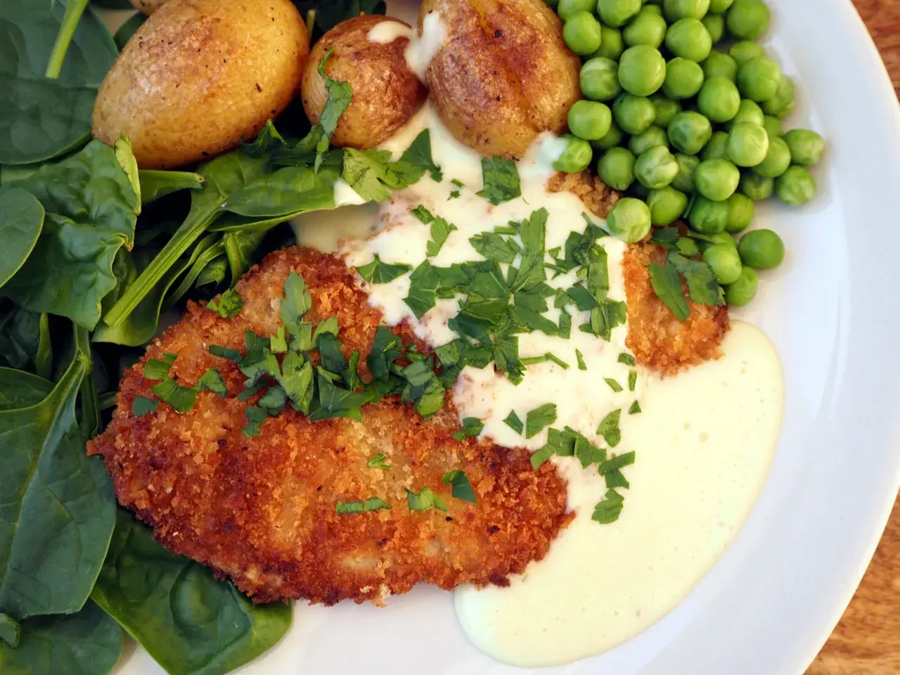 Krispig schnitzel med krämig sås till schnitzel, serverad med små potatisar, gröna ärtor och färsk spenat.