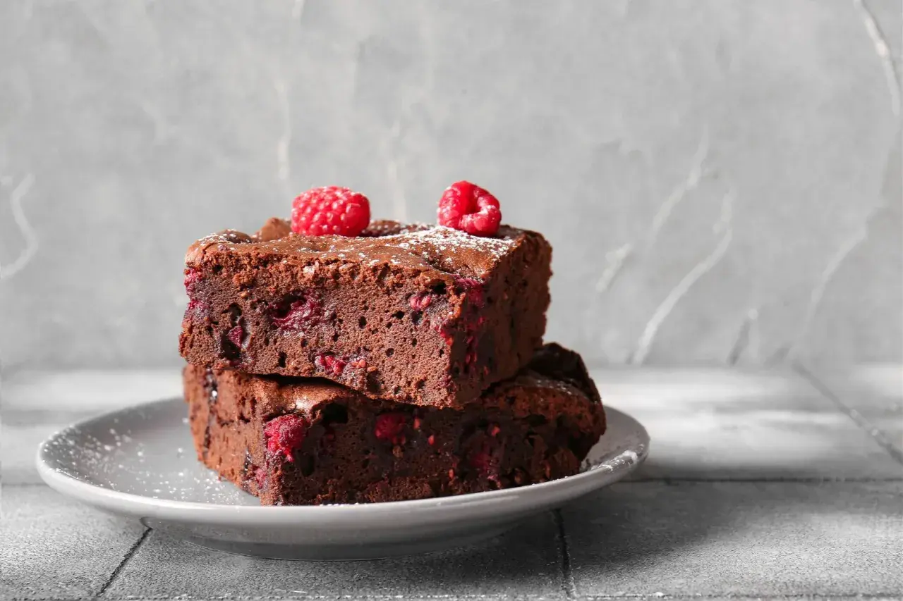 Dwa wilgotne brownie z malinami, posypane cukrem pudrem, na szarym talerzyku. Idealne na deser.