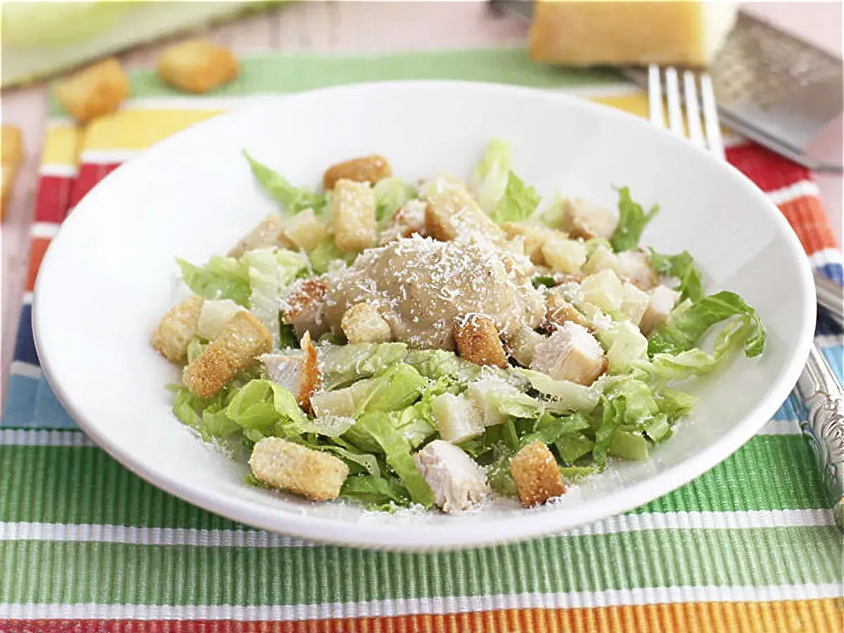 Una deliciosa ensalada cesar con pollo, crutones crujientes y queso parmesano rallado, servida en un plato blanco sobre un mantel colorido.