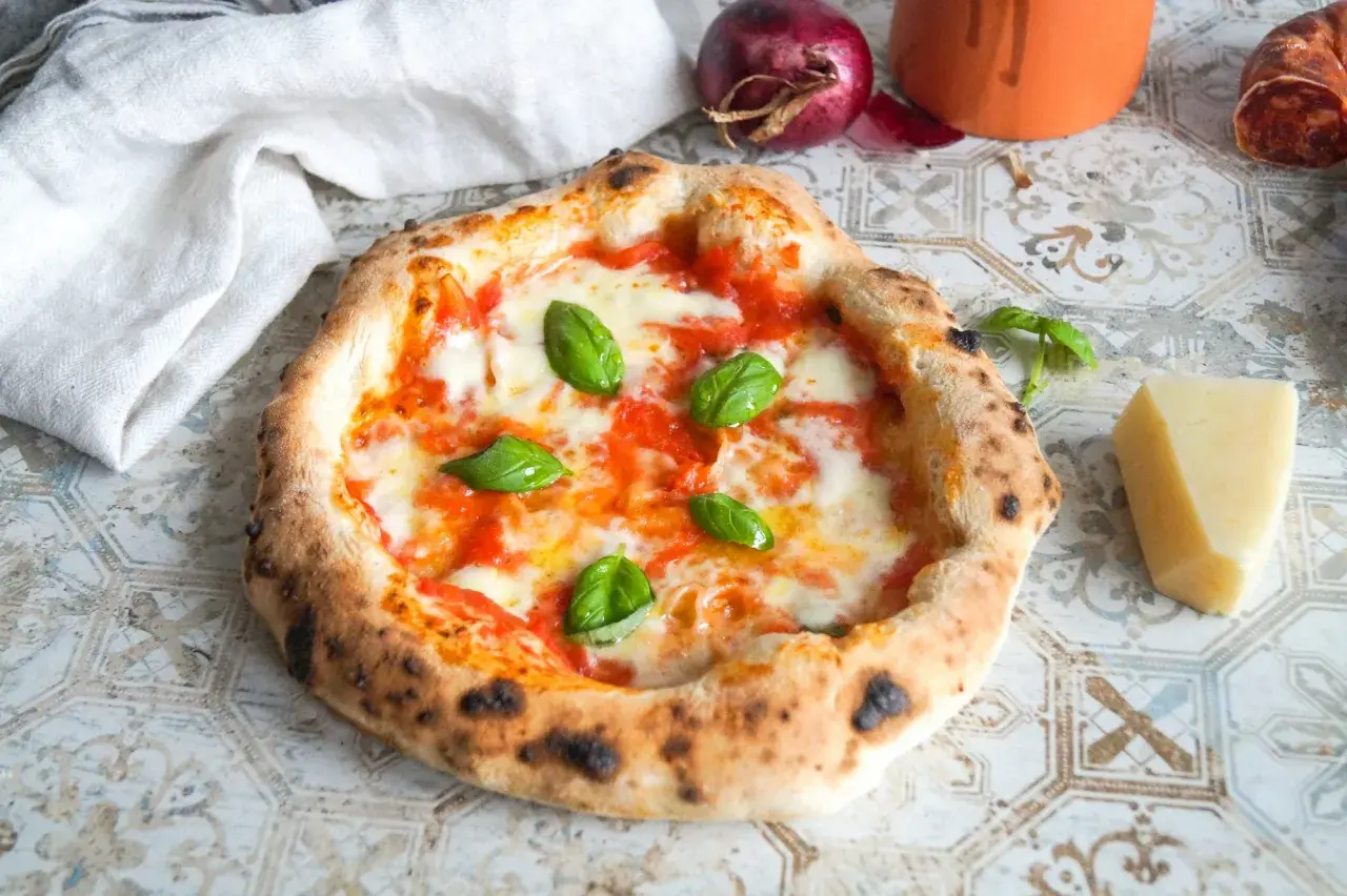 Eine authentische Pizza Napoletana mit frischem Basilikum, geschmolzenem Mozzarella und Tomatensauce. Daneben ein Stück Parmesan und eine rote Zwiebel.