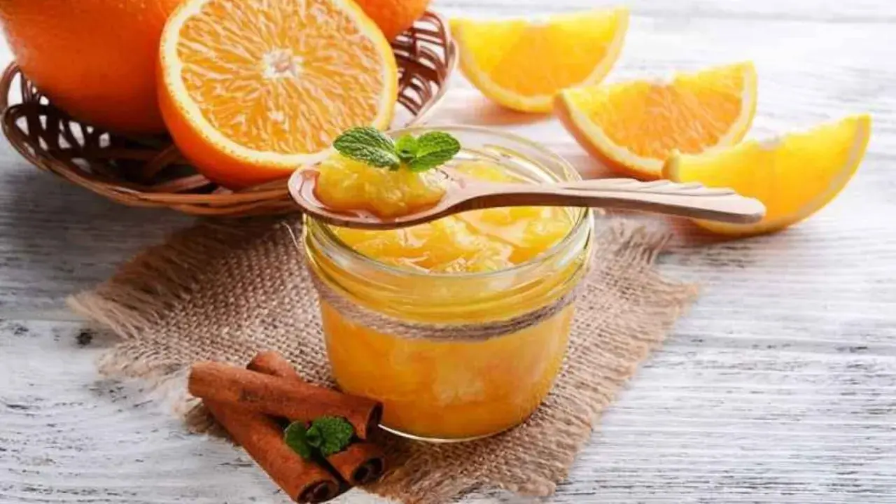 Tarro de mermelada de naranja casera, con naranjas frescas y canela en rama. Una cucharada muestra la textura.