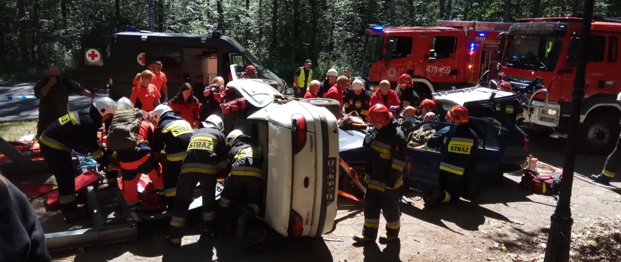 Akcja ratunkowa ksrg po wypadku. Strażacy wyciągają poszkodowanych z przewróconego samochodu. W tle wozy strażackie i karetka.