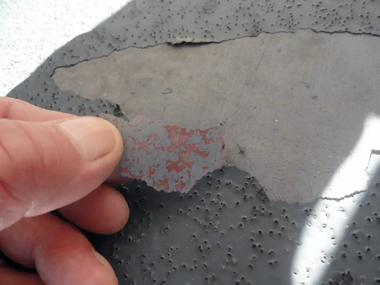 Dedo retirando capa desprendida de pintura gris sobre hormigón con picaduras, indicando necesidad de reparación de hormigón.