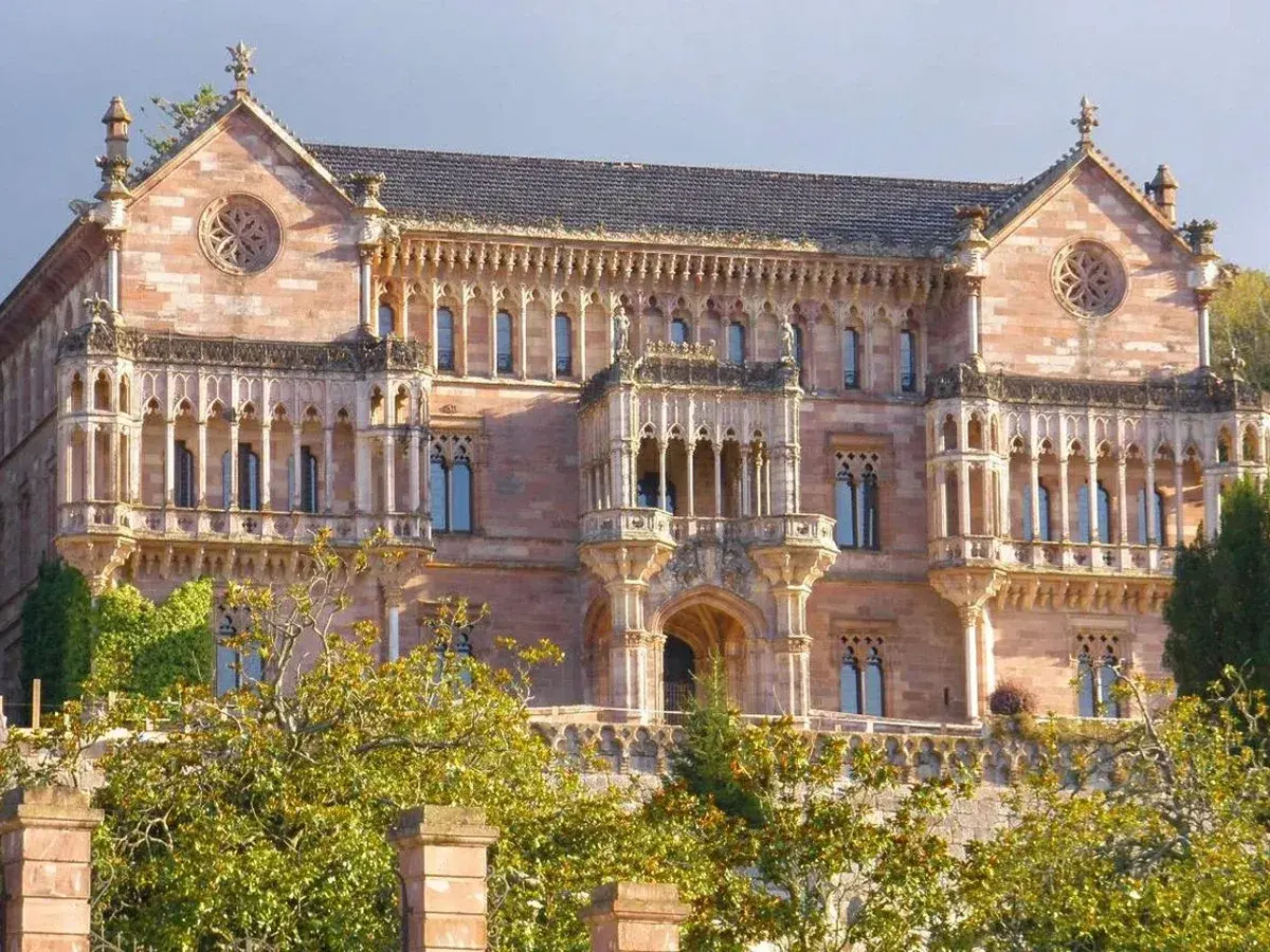 El **Palacio de Comillas**, una joya arquitectónica de piedra rosada, se alza majestuoso entre la vegetación, con sus balcones y rosetones detallados.