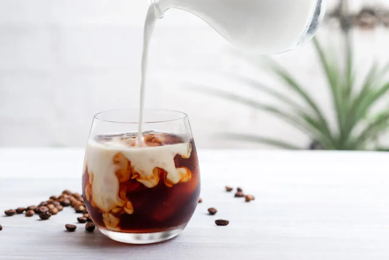 Mjölk hälls i ett glas med iskaffe, perfekt för ett enkelt iskaffe recept. Kaffebönor ligger utspridda bredvid.
