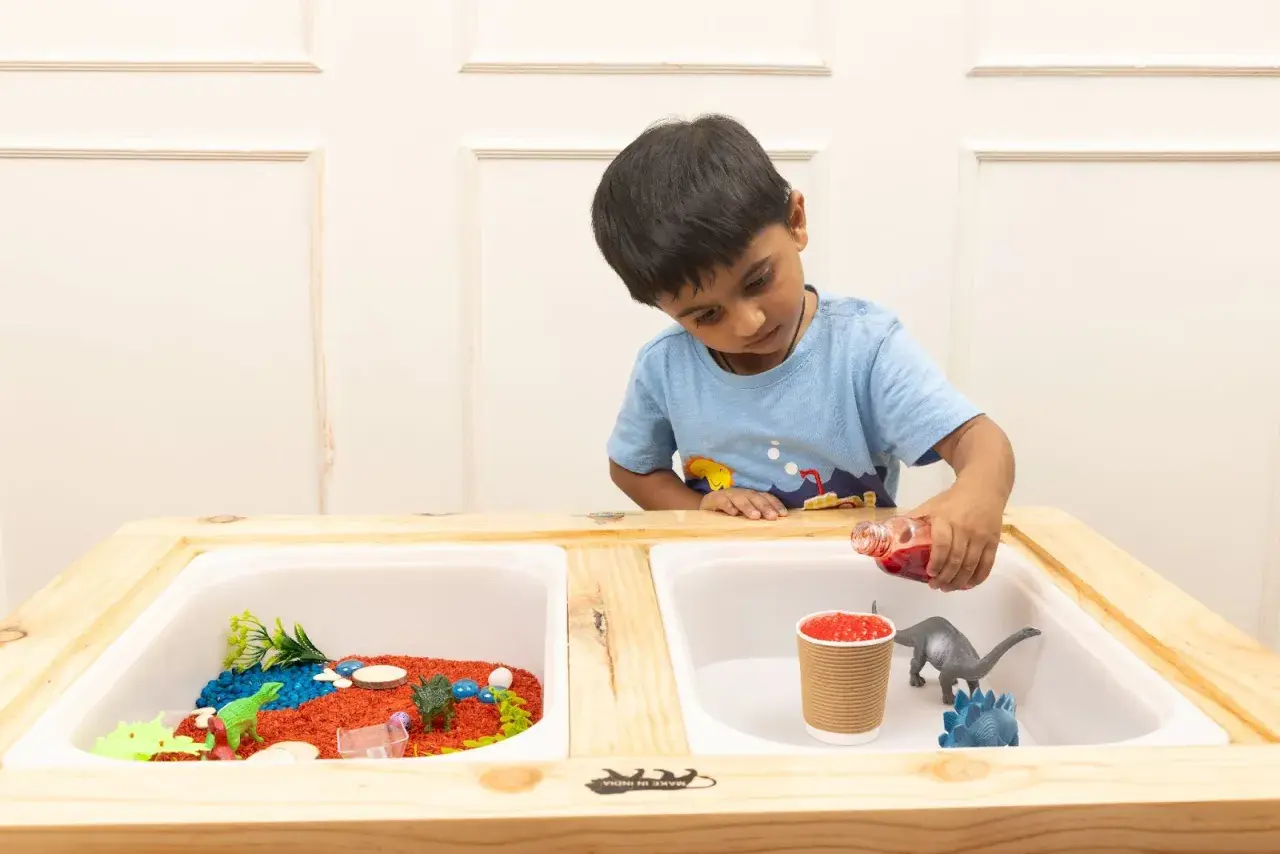 A boy pours red liquid into a sensory table with dinosaurs and colorful rice, exploring fun sensory table ideas.