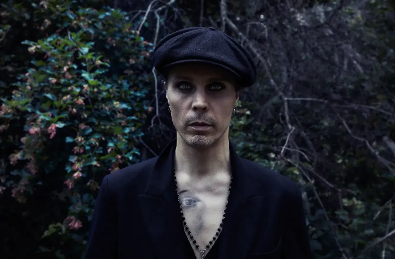 Ville Valo w czarnym kaszkiecie i marynarce, z intensywnym makijażem oczu, stoi na tle ciemnej, leśnej roślinności.