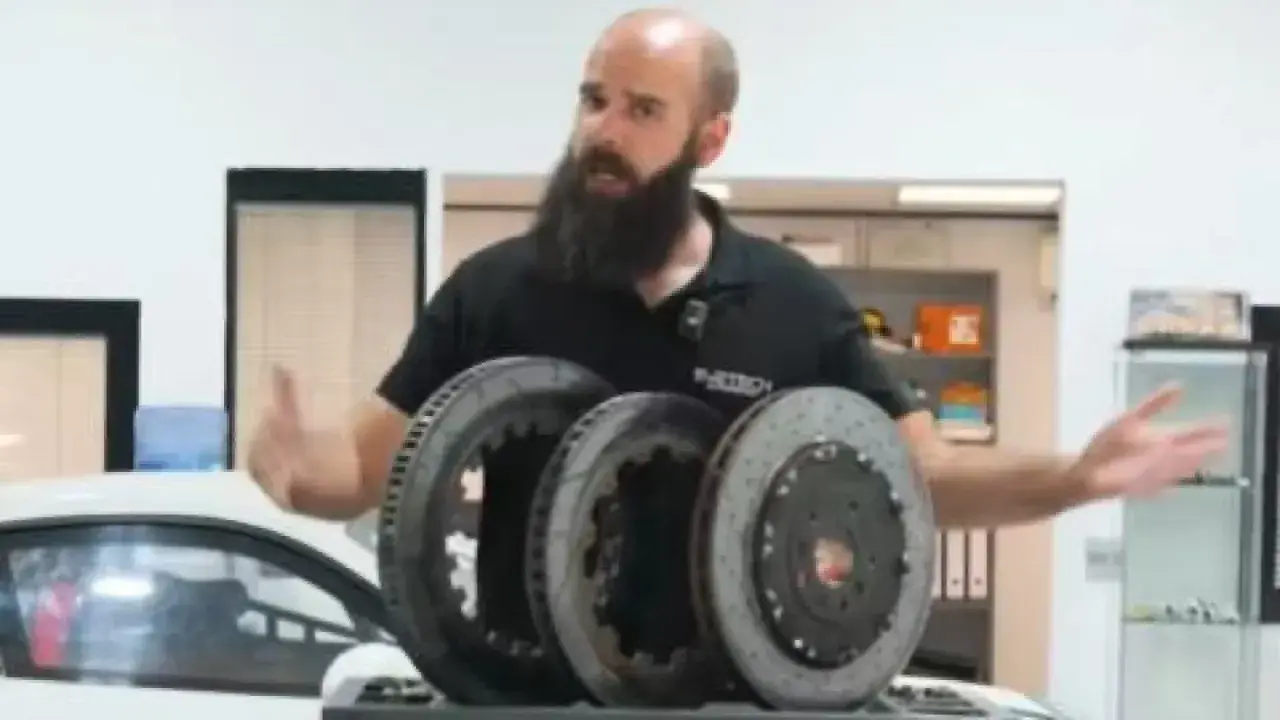 Hombre con barba muestra tres discos de freno, uno de ellos con perforaciones, para mejorar la refrigeración en un coche deportivo.