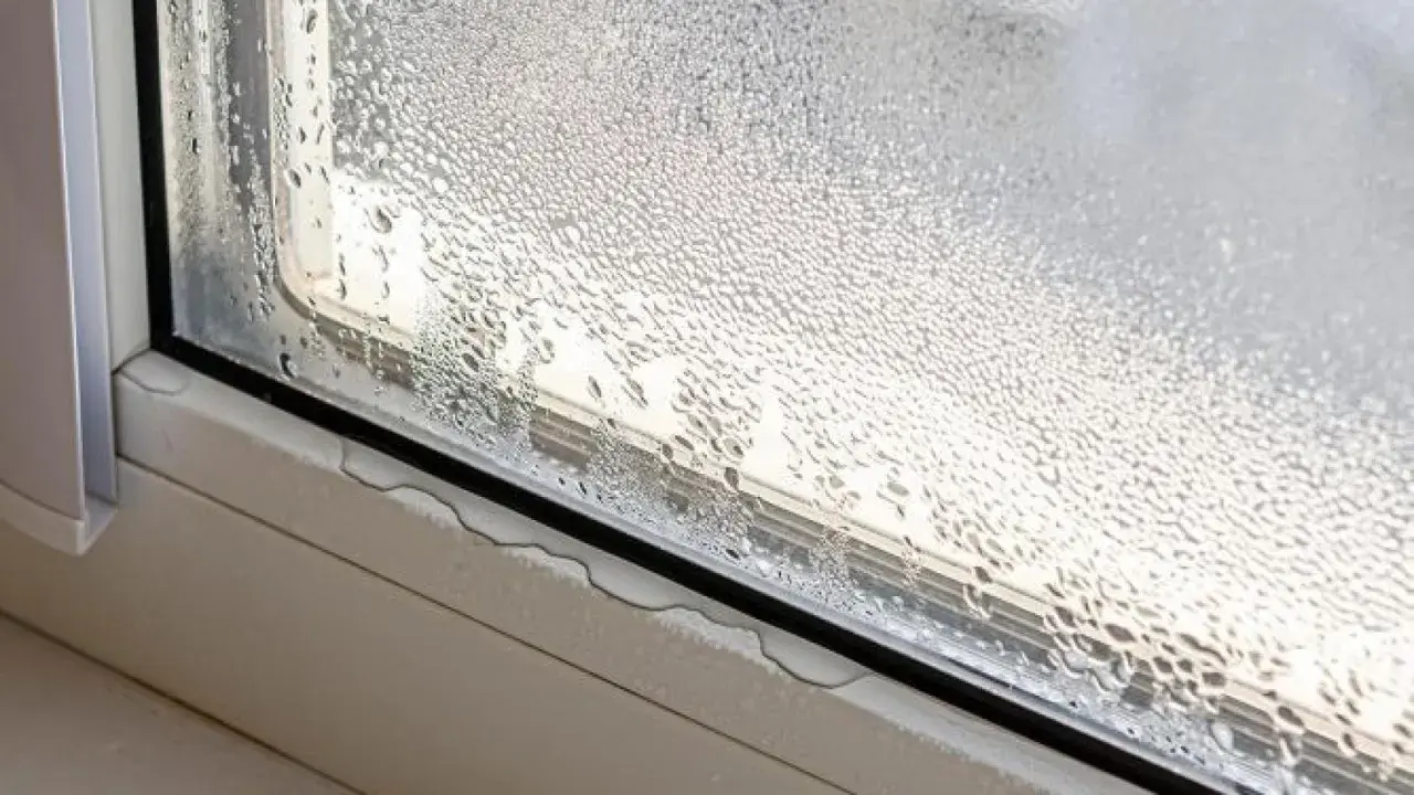 Gotas de agua cubren el cristal de la ventana, indicando condensación. El alféizar muestra pequeñas acumulaciones de agua.