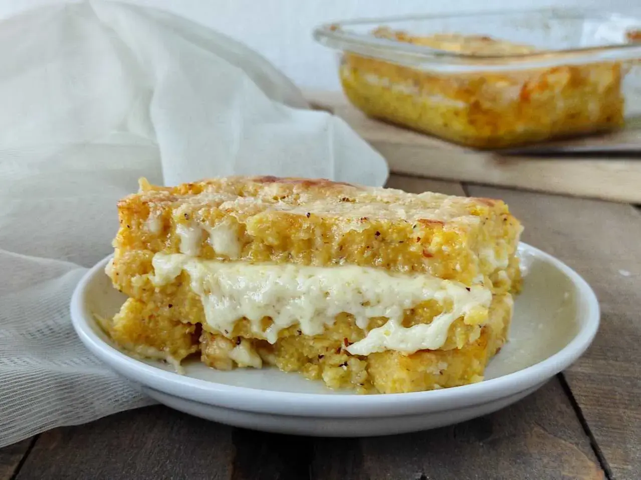 Una fetta di pasticcio di polenta con formaggio filante, cotto al forno e servito su un piatto bianco.