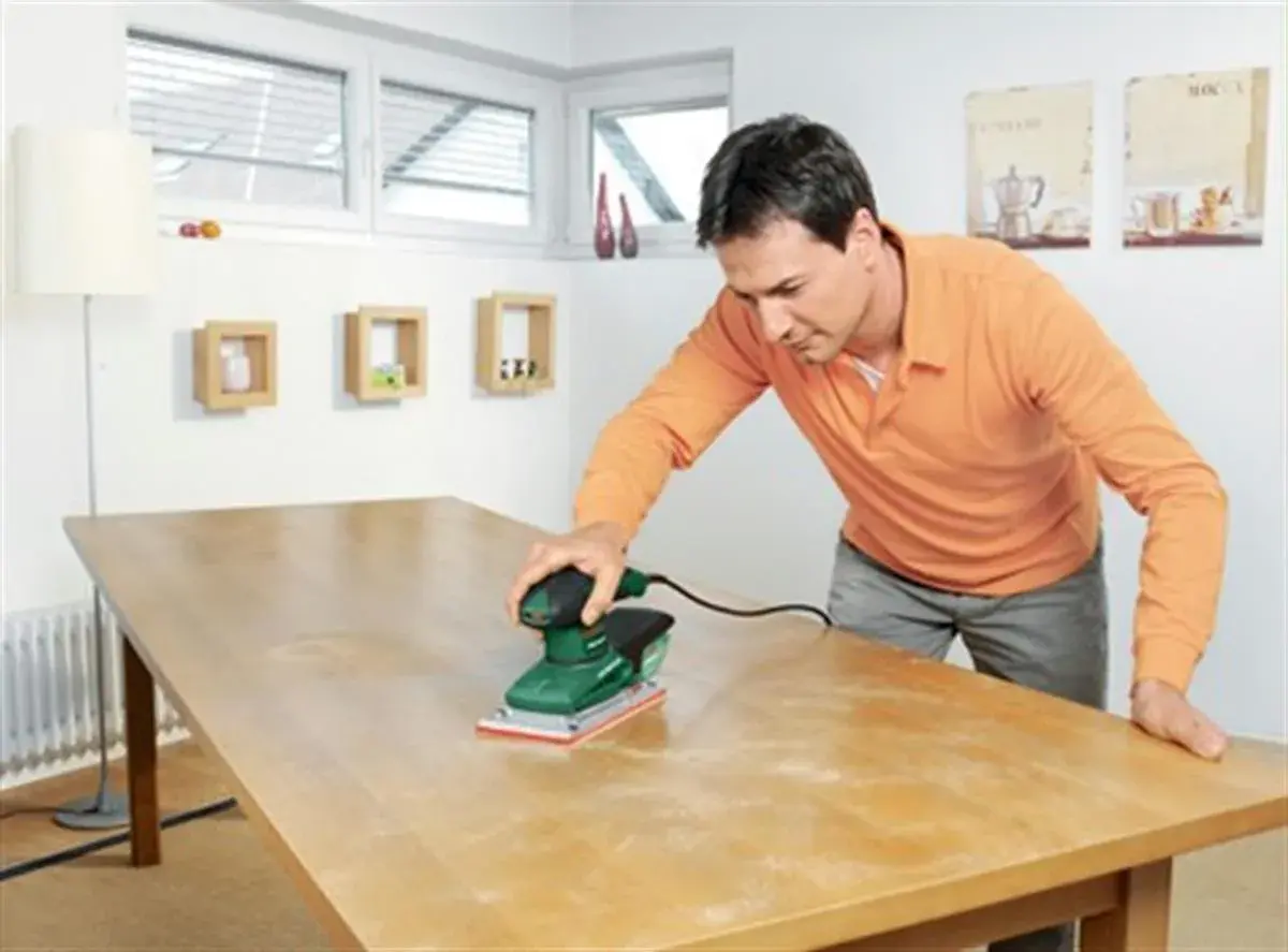 Un homme utilise une ponceuse vibrante ou excentrique pour rénover une table en bois.