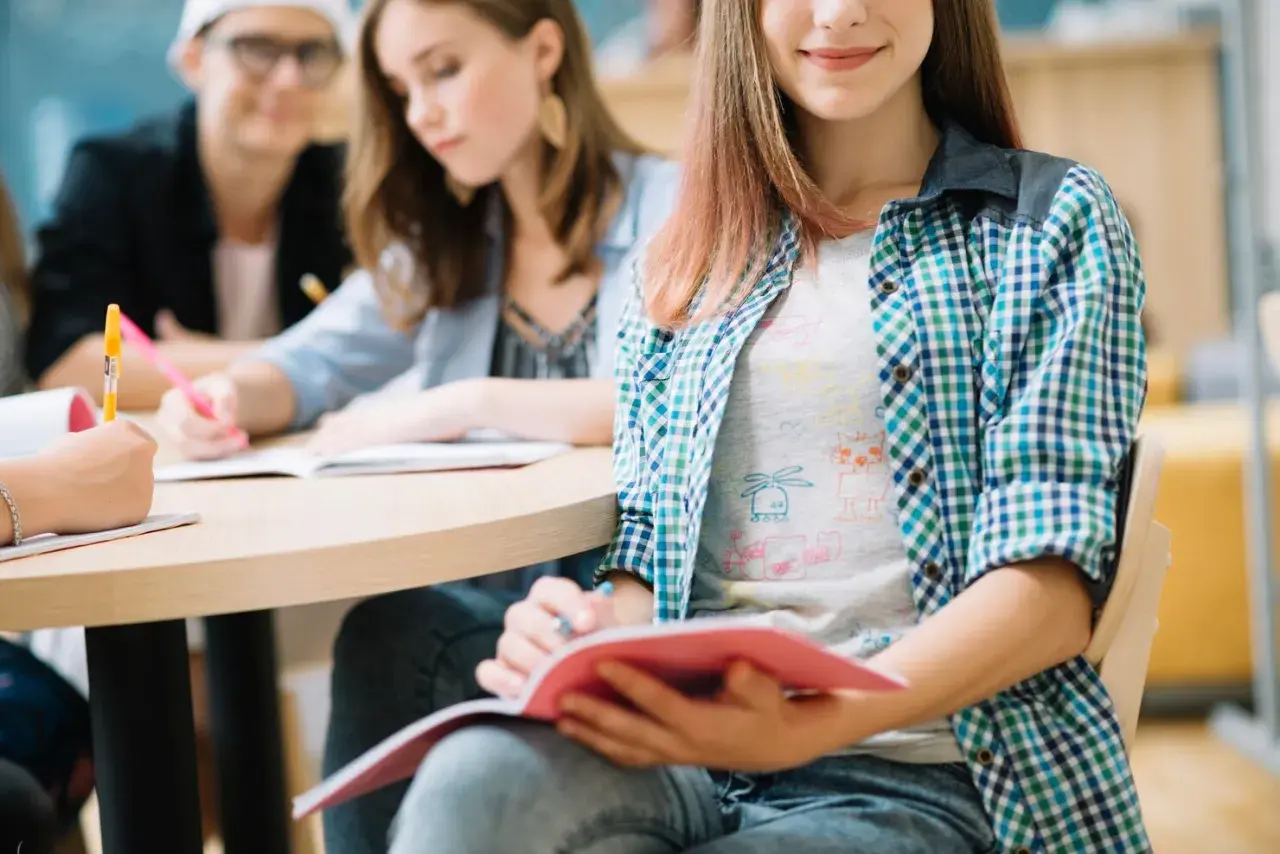 Uczniowie przy stole, skupieni na nauce biologii. Jedna z dziewczyn notuje, druga czyta, trzecia uśmiecha się, trzymając zeszyt.
