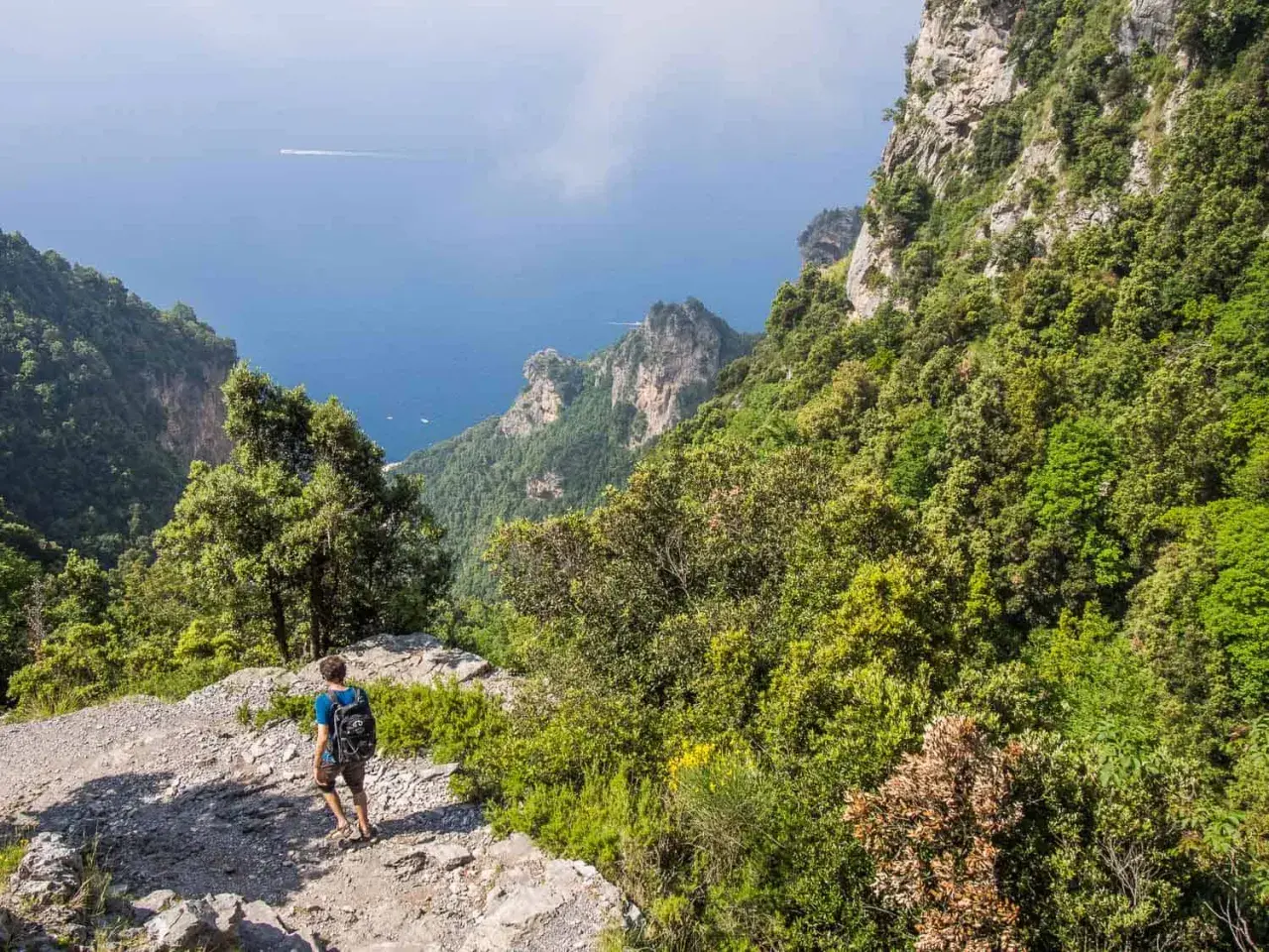 A hiker walks along the rocky path of the Sentiero degli Dei, with lush green mountains and the blue sea stretching out below.