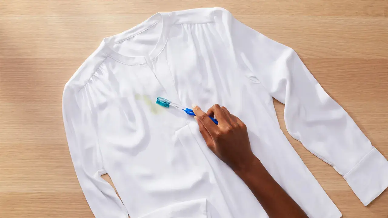 Une main utilise une brosse à dents pour enlever une tache verte sur une chemise blanche. Astuce : comment enlever de la colle sur un vêtement.