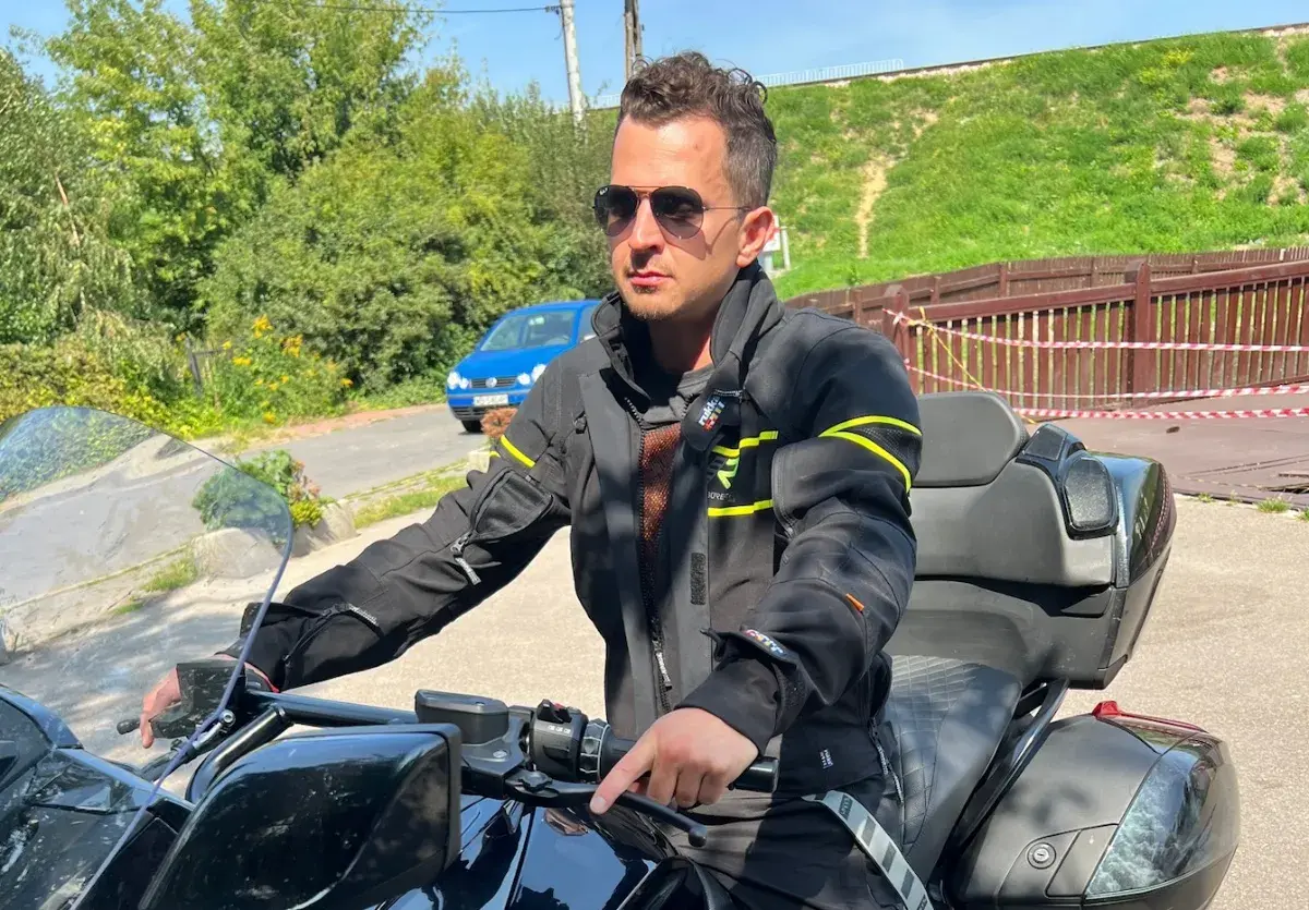 Mężczyzna w okularach przeciwsłonecznych i czarnej kurtce motocyklowej, gotowy na podróż.