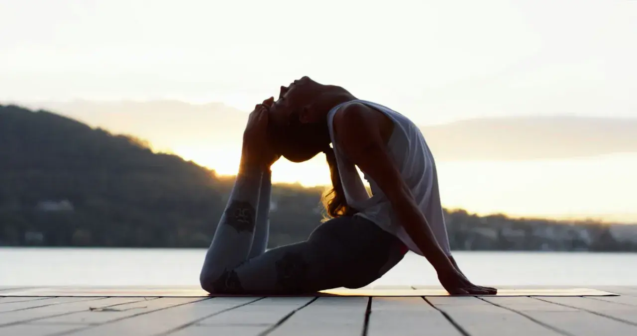 Silhouette d'une femme en posture de yoga au bord de l'eau, le soleil se levant derrière elle. Ses leggings sont ornés de symboles, évoquant les mantra.