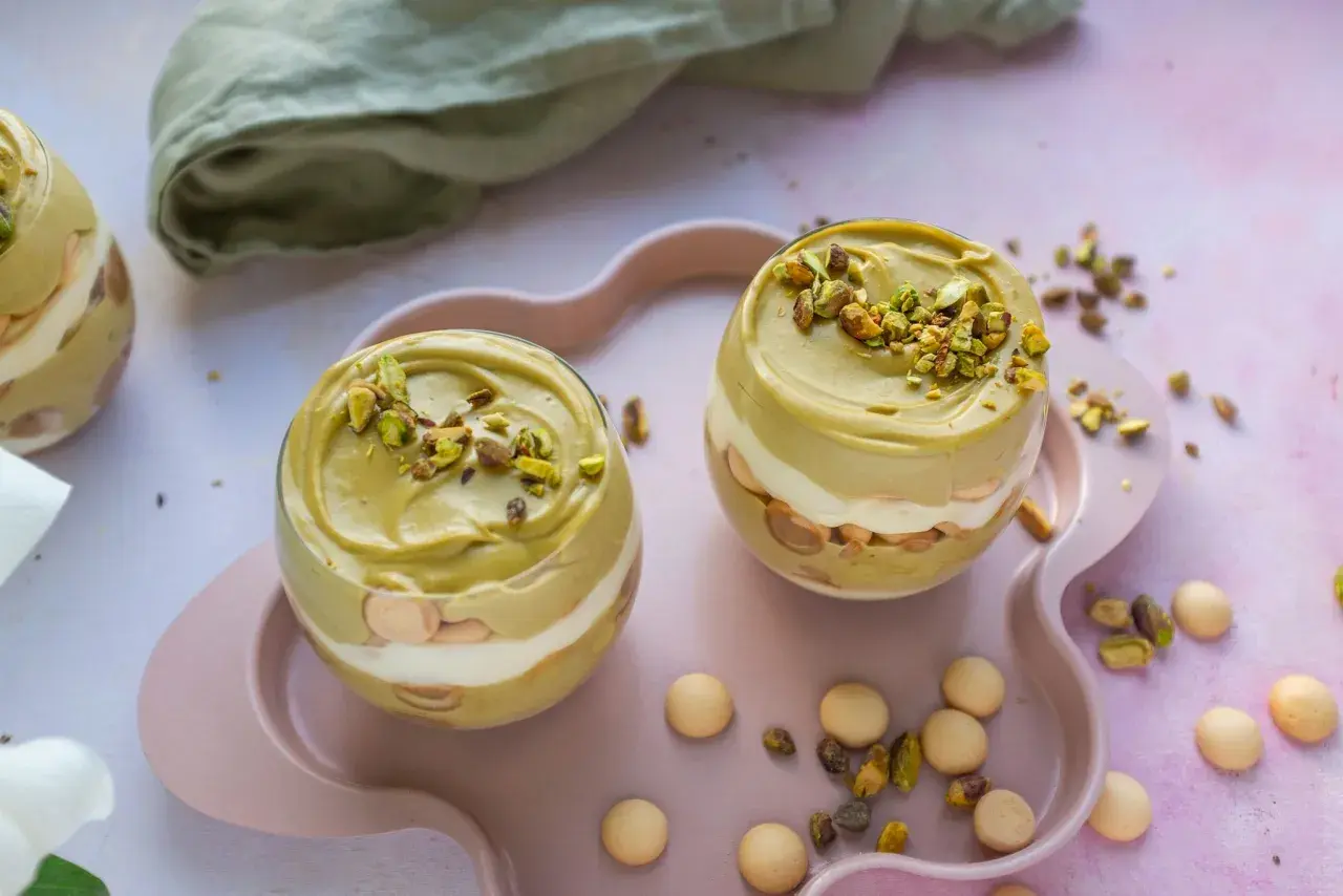 Dwa pucharki z deserem: krem pistacjowy z mascarpone, przełożony biszkoptami i bitą śmietaną, posypany pistacjami.