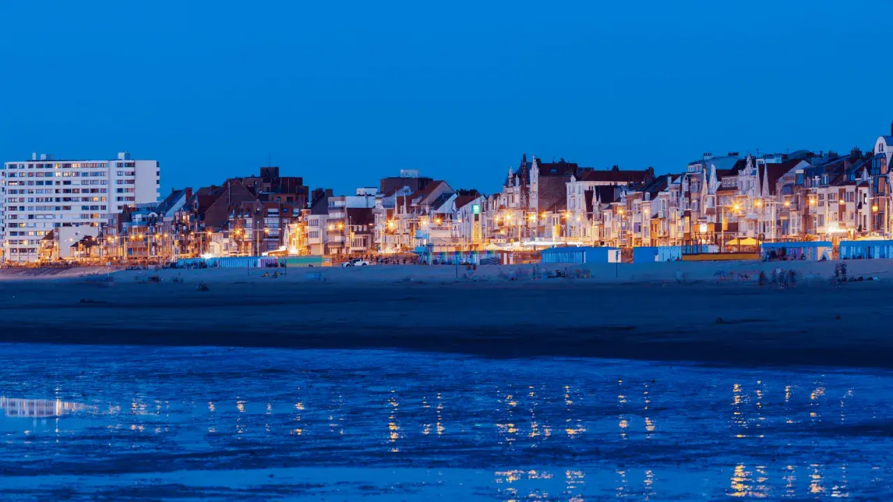 Kvällsljus över strandpromenaden i Dunkirk stad. Husen längs kusten lyser upp mot den mörka himlen, med reflektioner i det lugna vattnet.